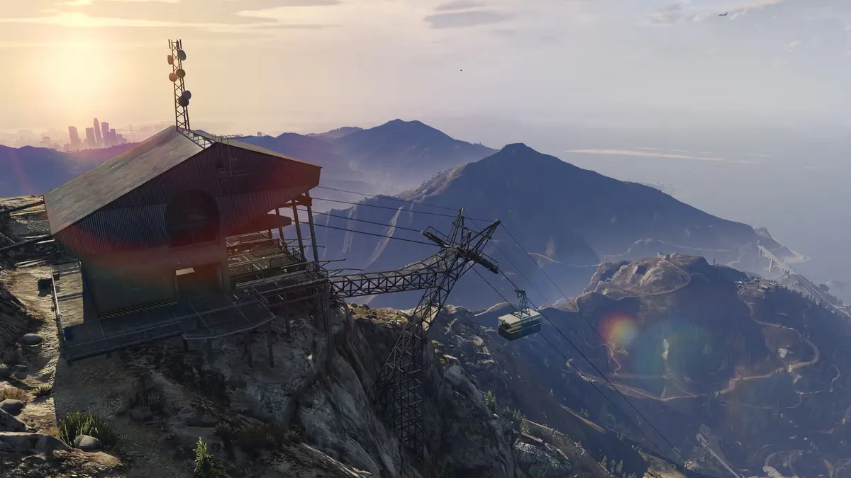 Capture de GTA 5 : station de téléphérique au sommet du mont Chiliad au lever du soleil, avec vue panoramique sur les montagnes, l'océan et la silhouette lointaine de Los Santos