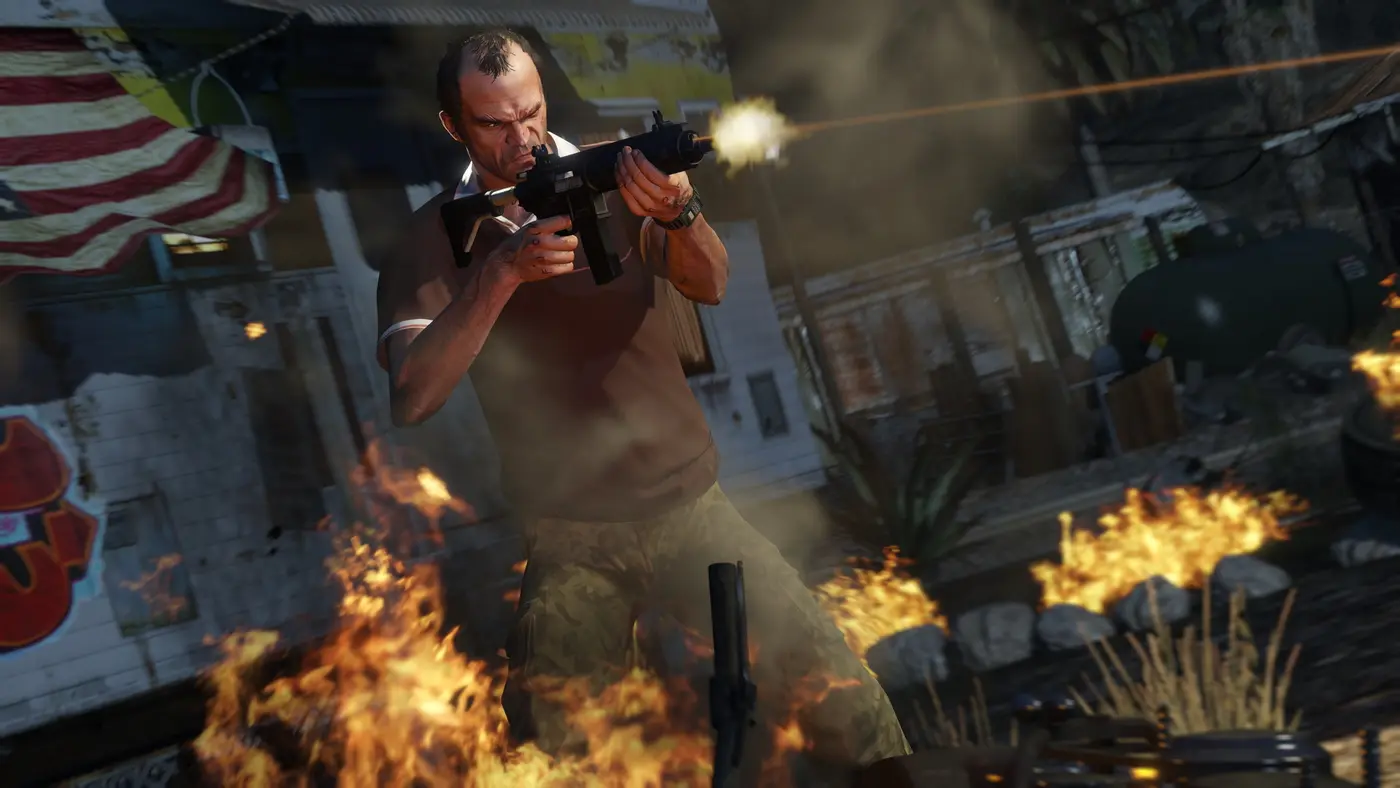 Trevor Philips tirant avec un pistolet-mitrailleur au milieu des flammes et de la fumée, dans une cour délabrée, un drapeau américain accroché au mur derrière lui