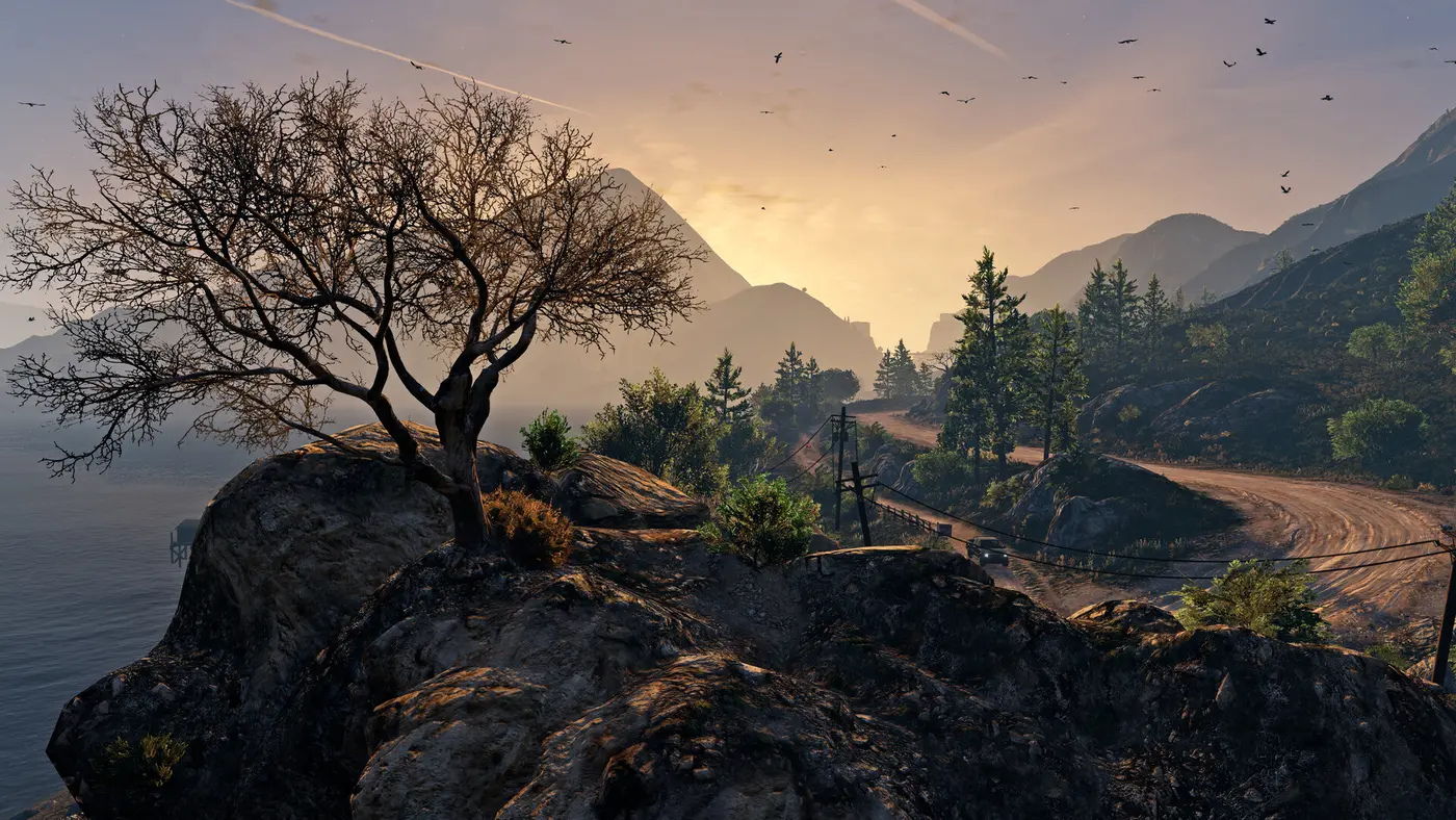 Capture officielle de GTA V : un arbre sec sur un rocher au bord de l'eau, une piste de terre qui serpente entre les pins et les montagnes au coucher du soleil