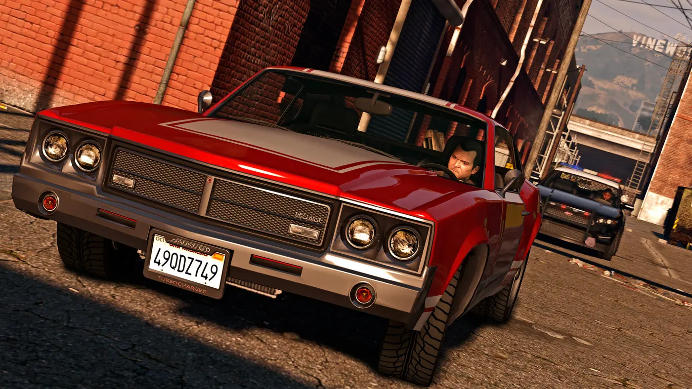 Capture officielle de GTA V : Michael au volant d'une Declasse Sabre Turbo rouge dans une ruelle de briques, une voiture de police derrière lui et le panneau Vinewood au loin