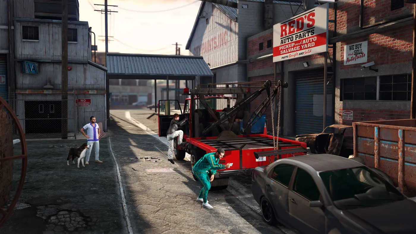 La devanture de Red's Auto Parts dans GTA Online, une casse automobile d'un quartier industriel de Los Santos au soleil couchant, une dépanneuse rouge garée devant l'enseigne, deux personnages en survêtement et un chien sur la chaussée