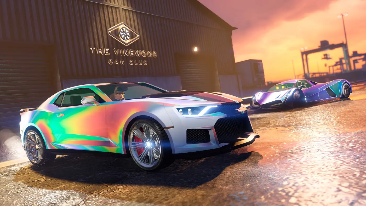 Capture de GTA Online : deux véhicules aux peintures caméléon irisées garés devant l'entrepôt du Vinewood Car Club au coucher du soleil sur Elysian Island, grues portuaires en arrière-plan