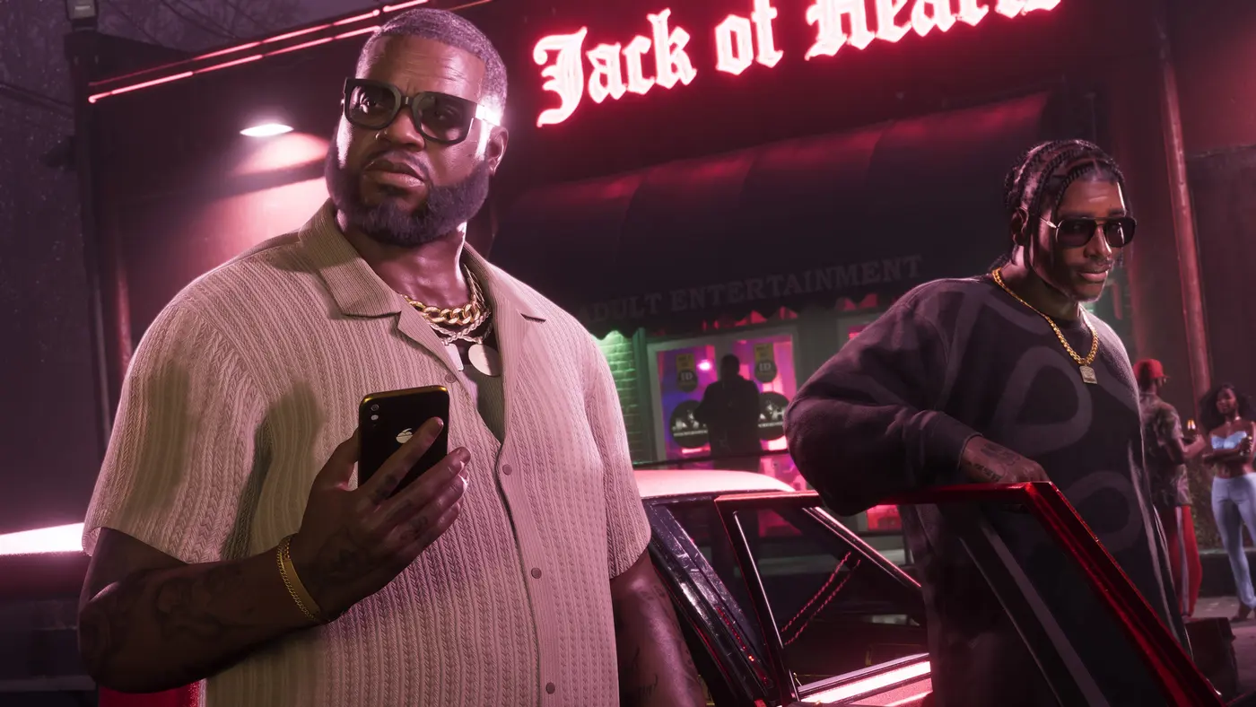 Un homme imposant en chemise claire et grosse chaîne en or tient un smartphone devant l'enseigne néon rouge Jack of Hearts d'un club pour adultes la nuit dans GTA VI, un second personnage à tresses près d'une voiture classique au capot rouge