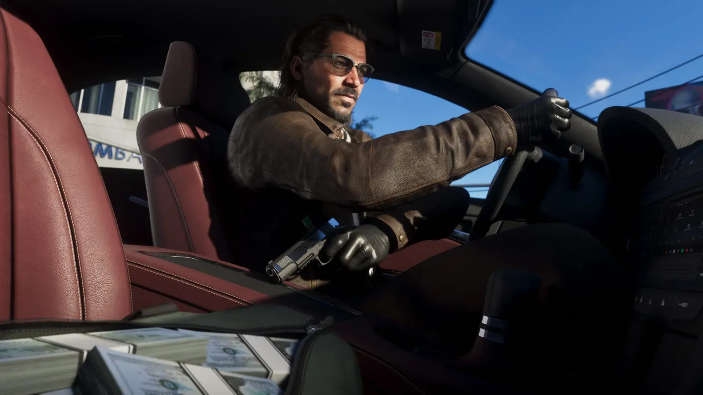 Raul Bautista dans GTA VI, homme à lunettes et catogan en veste de cuir et gants noirs, assis au volant d'une voiture, un pistolet et des liasses de billets posés près de lui en plein jour