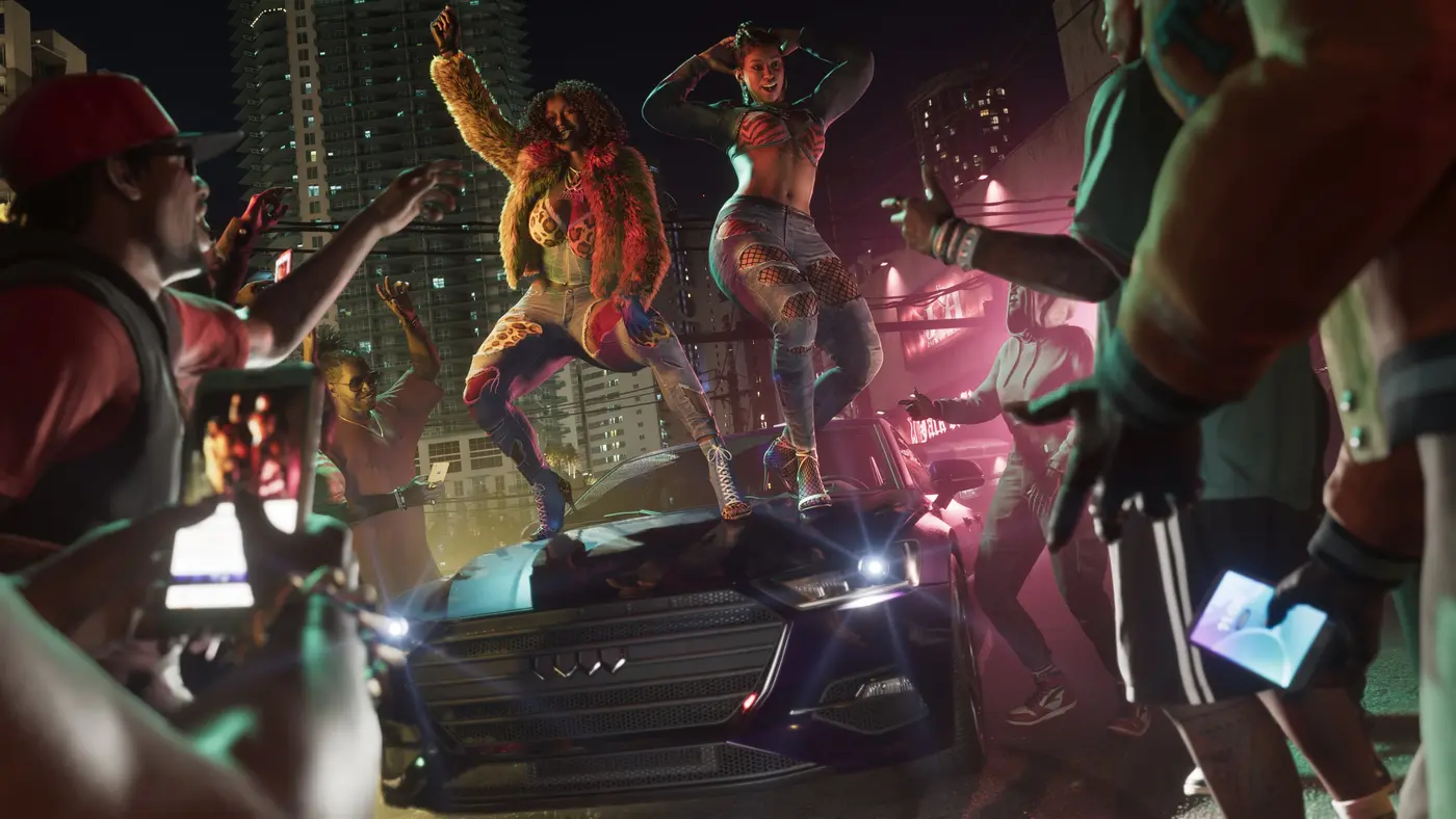 Le duo Real Dimez dans GTA VI, deux femmes dansant sur le capot d'une voiture lors d'un rassemblement nocturne en ville, une foule autour d'elles téléphone en main, gratte-ciel illuminés en arrière-plan