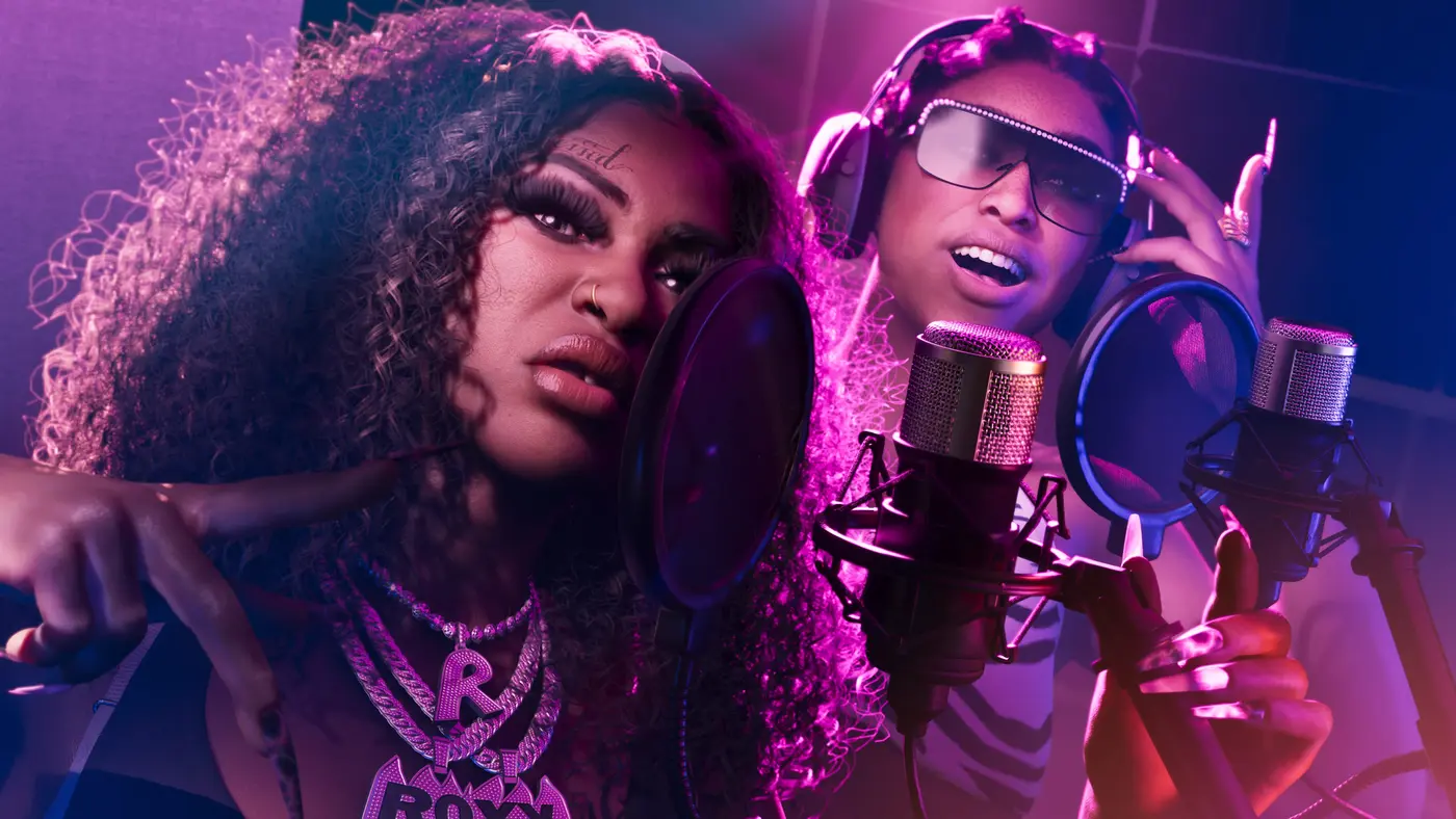 Deux artistes féminines de GTA VI enregistrent en studio, penchées vers des micros professionnels équipés de filtres anti-pop, casque sur les oreilles, lunettes pailletées et pendentif ROXY, baignées d'un éclairage néon rose et violet